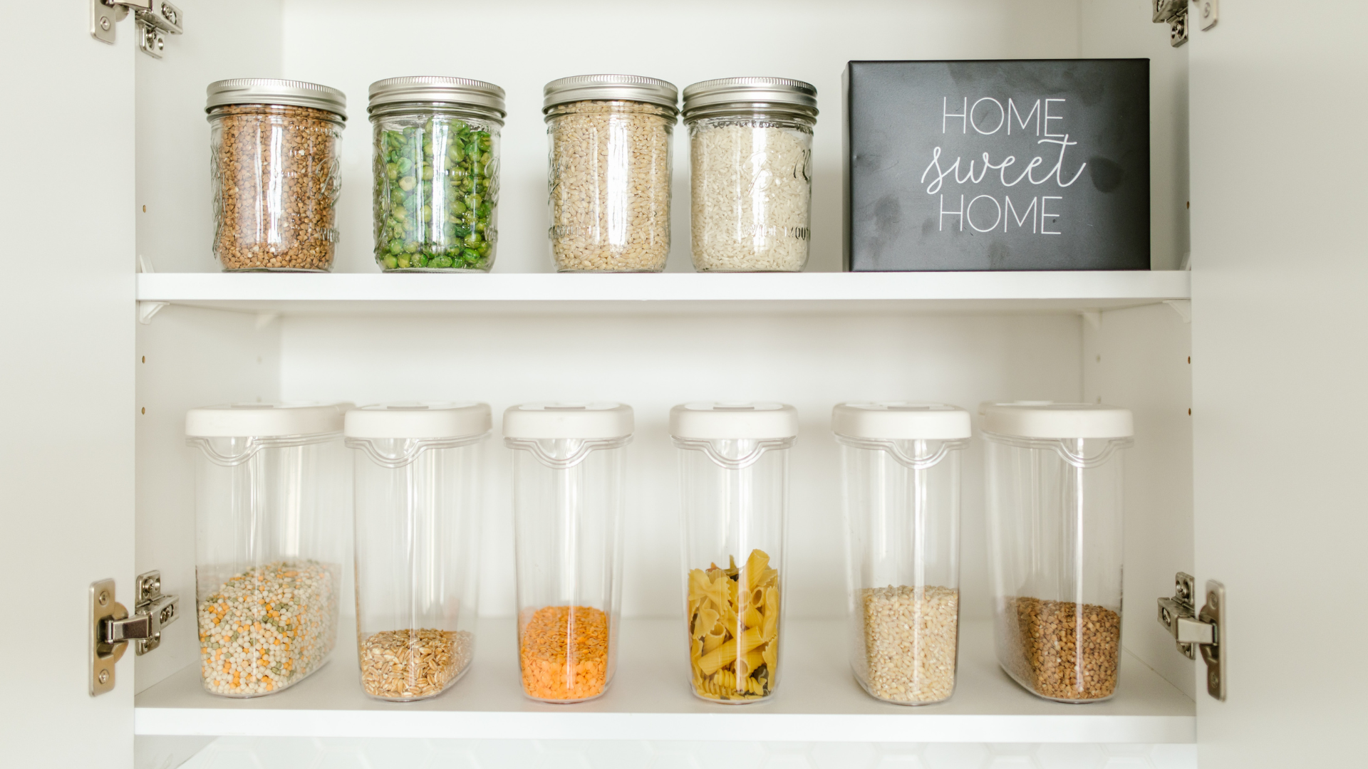Clear plastic pantry cereal organizers and clear glass food jars sit in white pantry cupboard