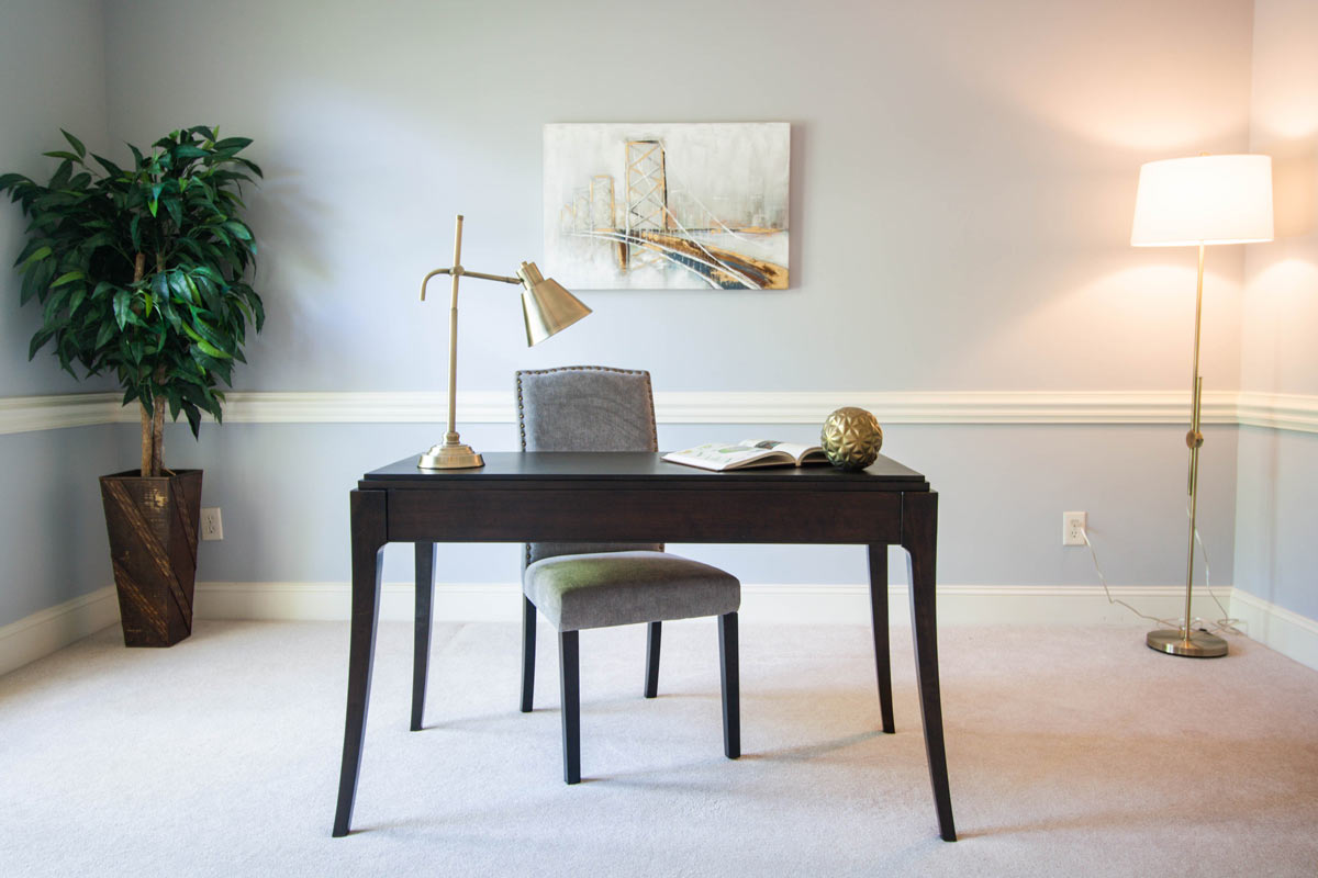 A vacant home staging of an office space with a traditional brown writing desk, accent lamps, wall art, and a faux tree