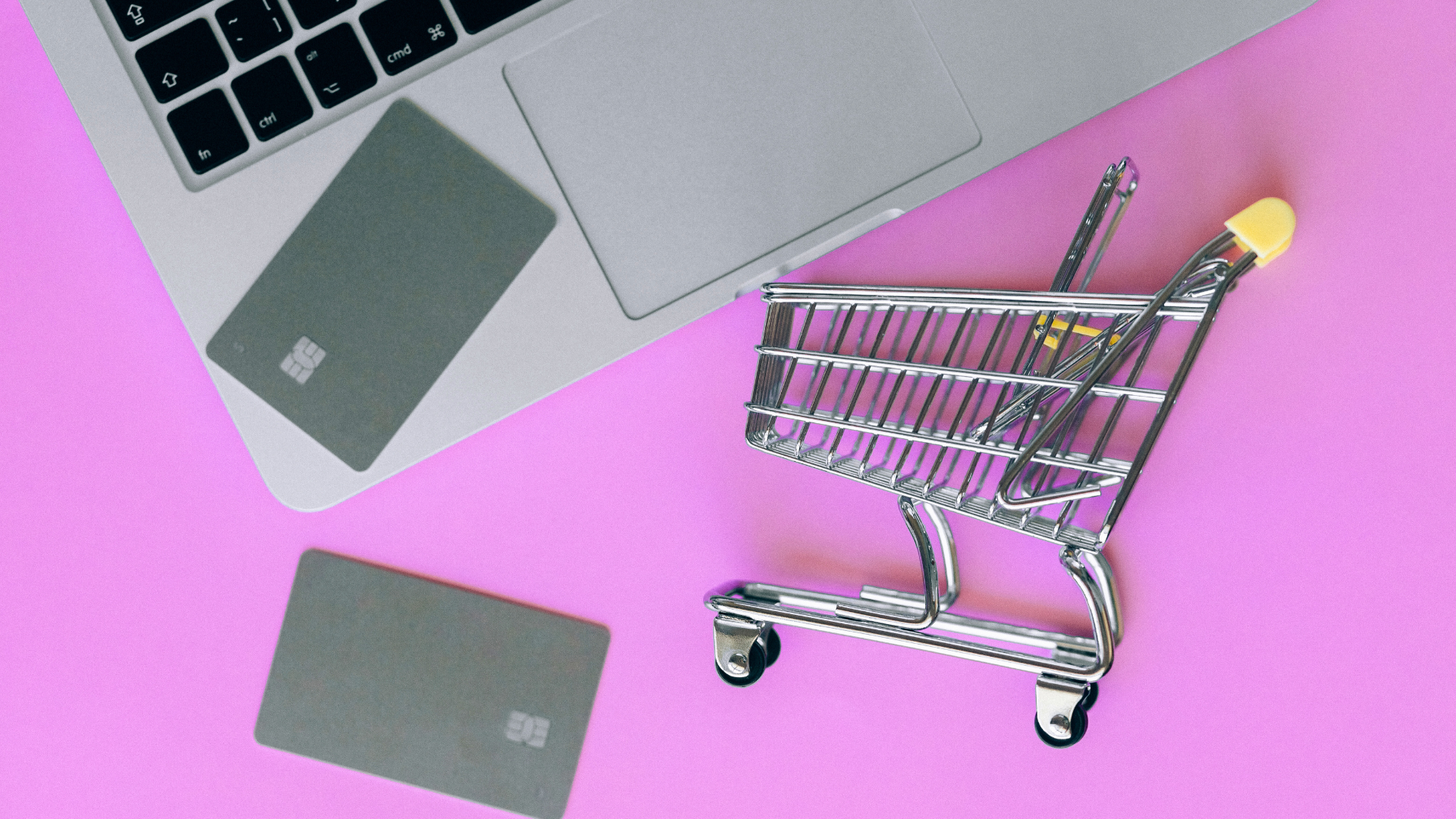 Silver shopping cart on pink surface with credit cards and laptop